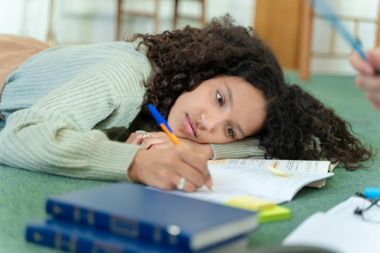 Teenage girl with curly hair lying on the floor resting her head on her arm while writing in a book, with textbooks and sticky notes beside her.