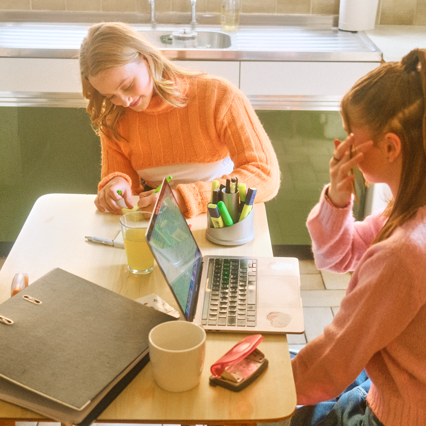 Two teenage girls sitting at a kitchen table with a laptop, folder, mug and pen pot in front of them, one writing and the other sitting beside her.