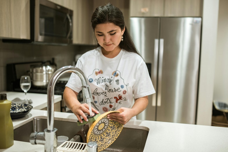 Teenage girl in a graphic T-shirt washing dishes at a kitchen sink, with a decorative plate in her hands and a fridge and hob in the background.