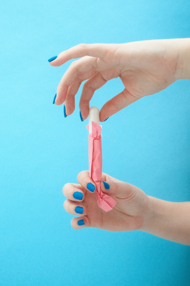 Two hands with blue nail polish hold an unwrapped tampon against a blue background.