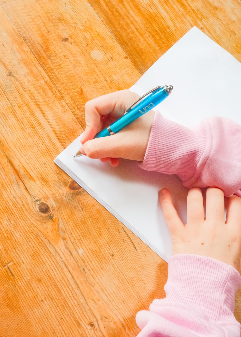 Overhead view of hands writing on a blank notebook page with a blue luna pen, on a wooden table.