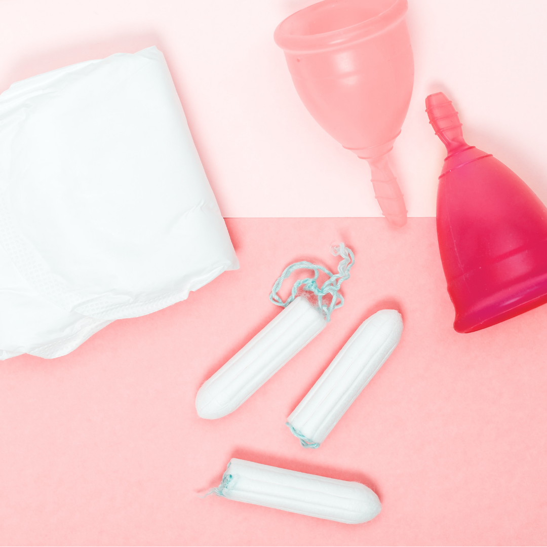 Flatlay of various period products against a pink background, including a period pad, a menstrual cup, a smaller pink menstrual cup, and three tampons with strings.