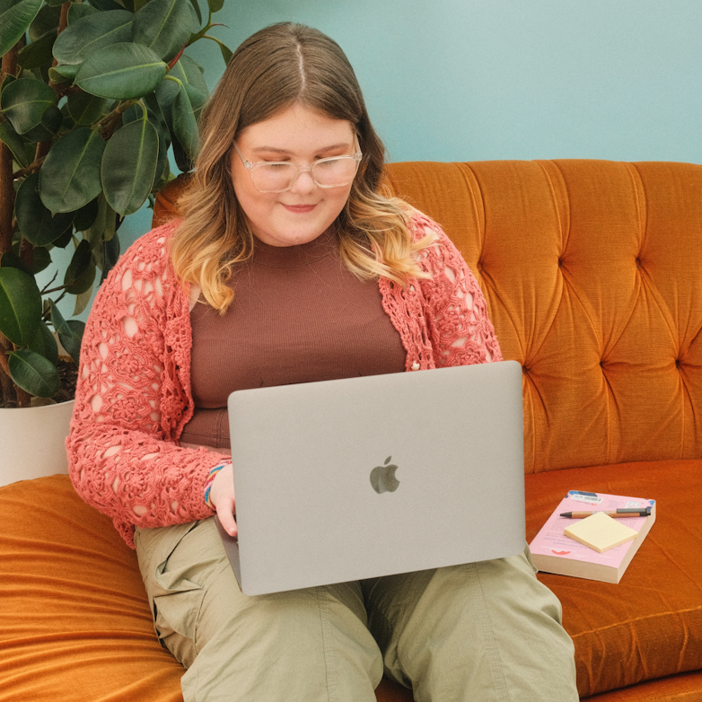 Teenage girl in a pink crochet cardigan sitting on an orange sofa smiling while using a laptop.