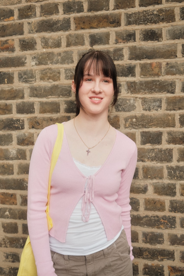 Teenage girl with dark hair and a fringe smiling in a pink cardigan against a brick wall.