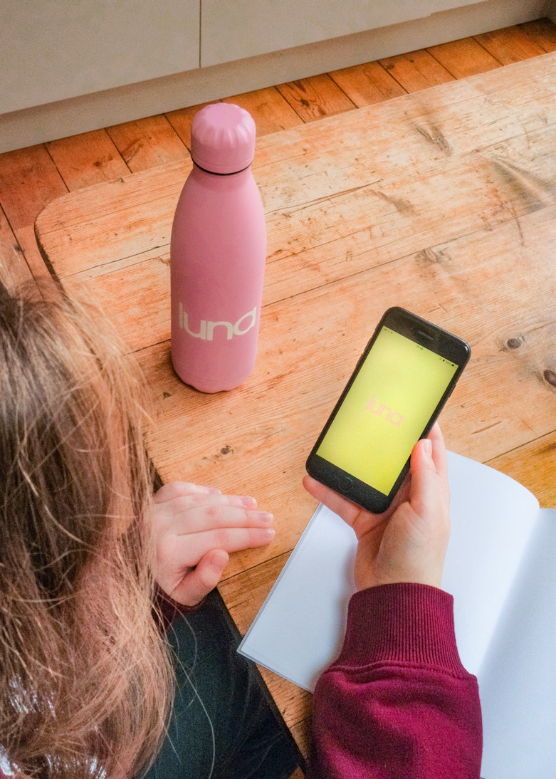 Teenage girl opening the luna app on her phone at a wooden desk next to a pink luna water bottle and a notebook.