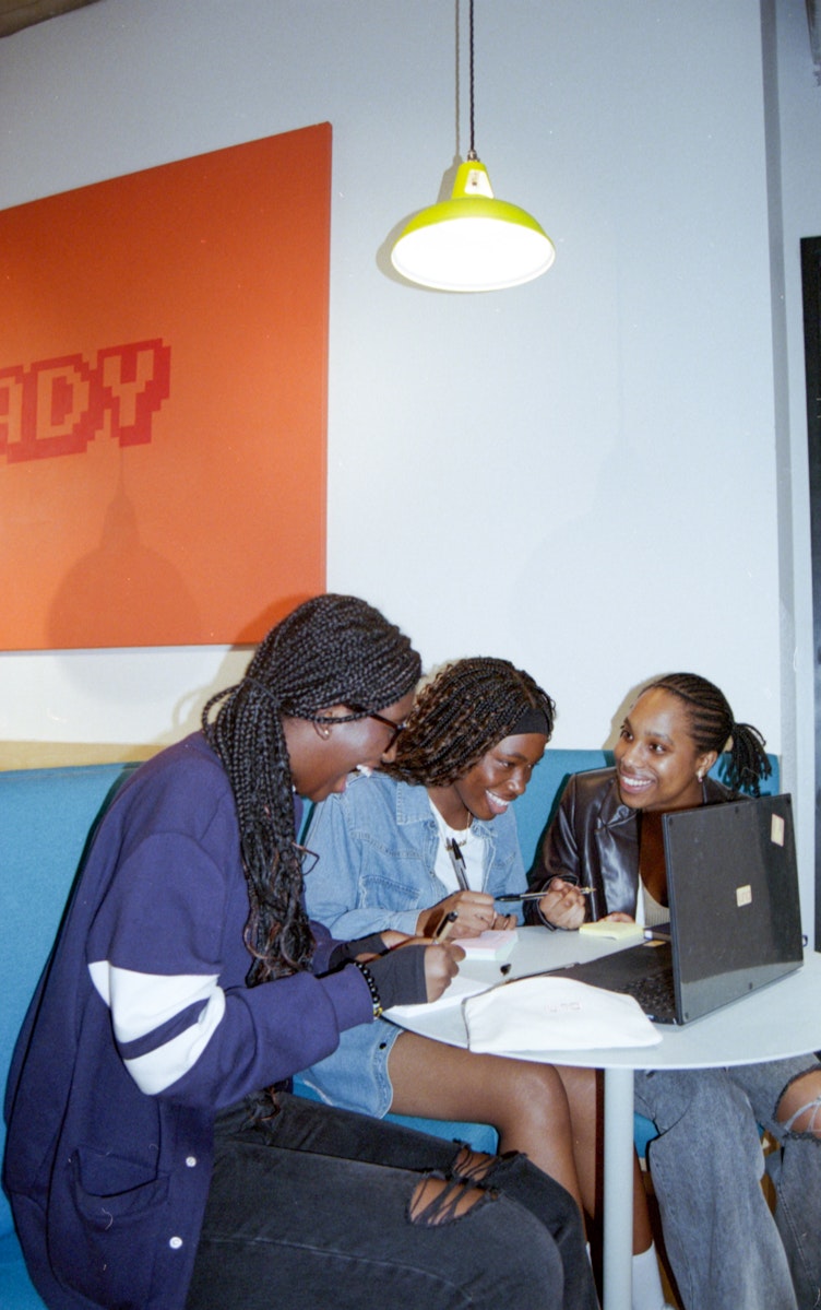 Three teenage girls sitting together at a table with a laptop, writing notes and smiling.