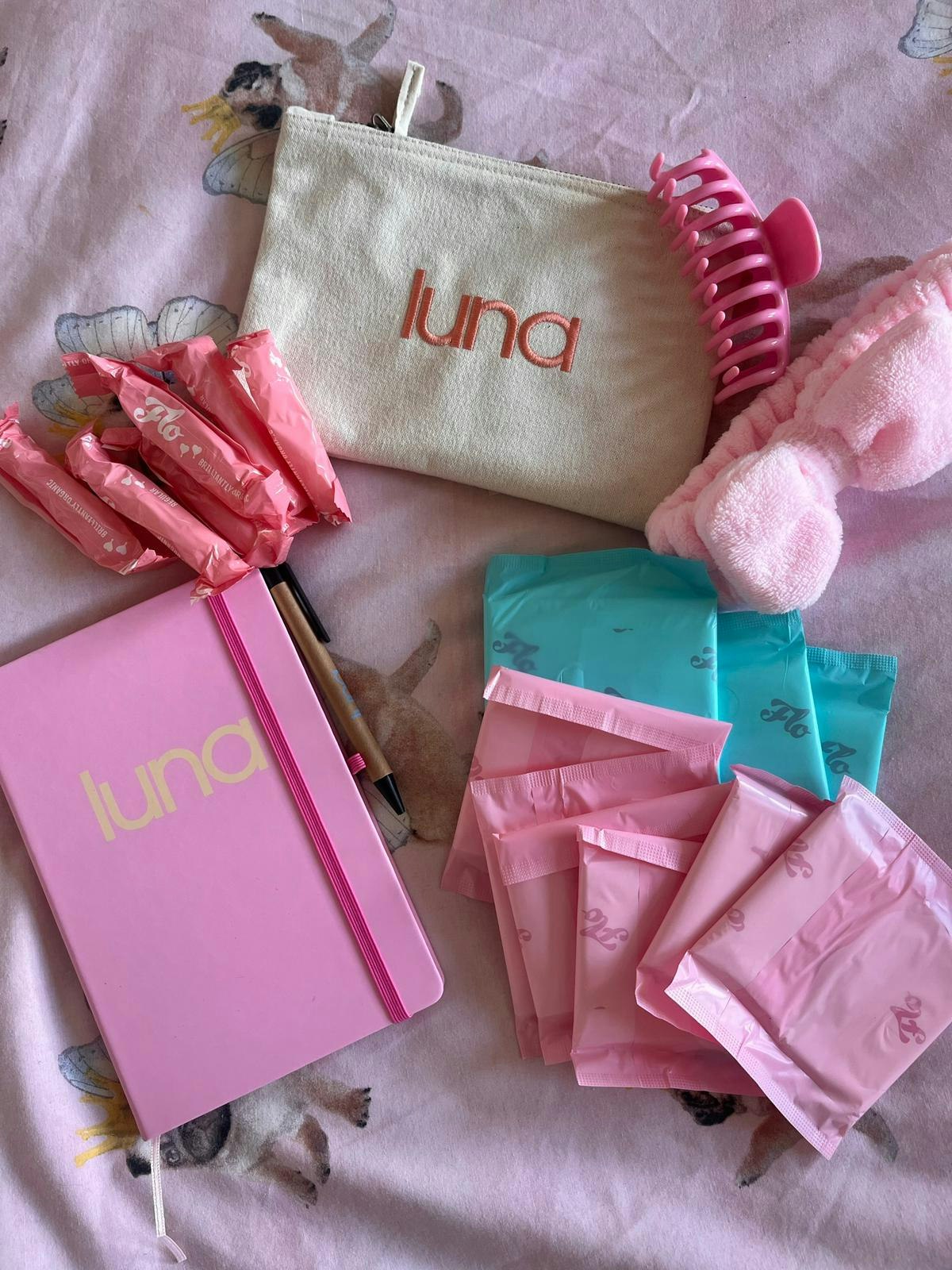 Flatlay of a luna period kit including individually wrapped period pads and tampons, a luna pouch, a luna notebook, a pink claw clip, and a fluffy headband.