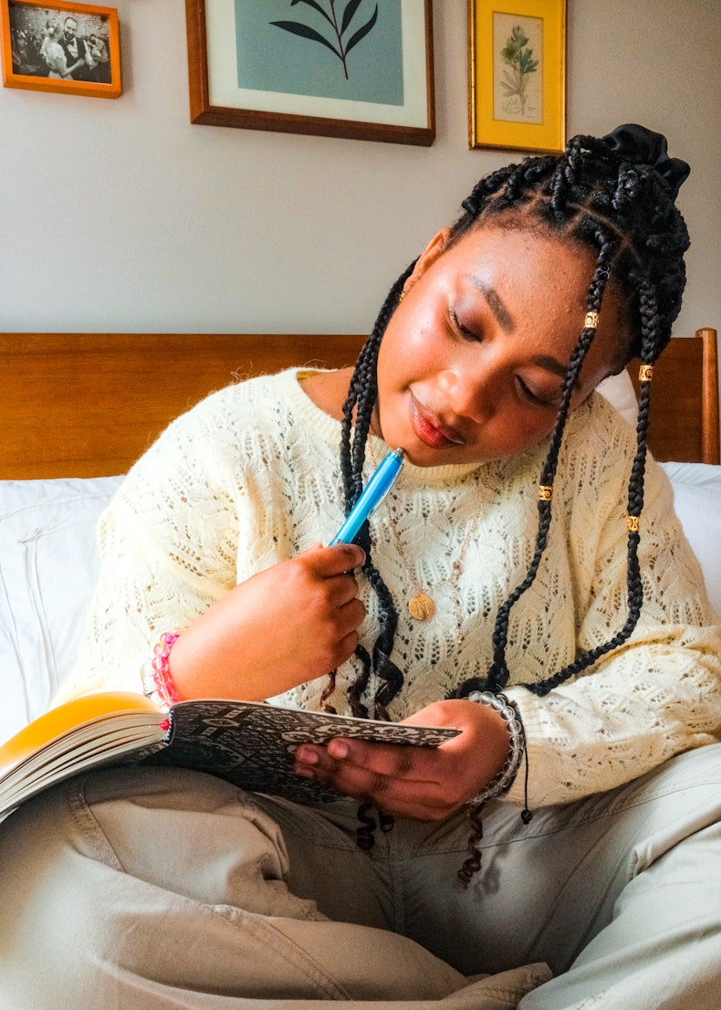 Teenage girl with braided hair sitting on a bed writing in a notebook with a blue pen, wearing a cream knit jumper.