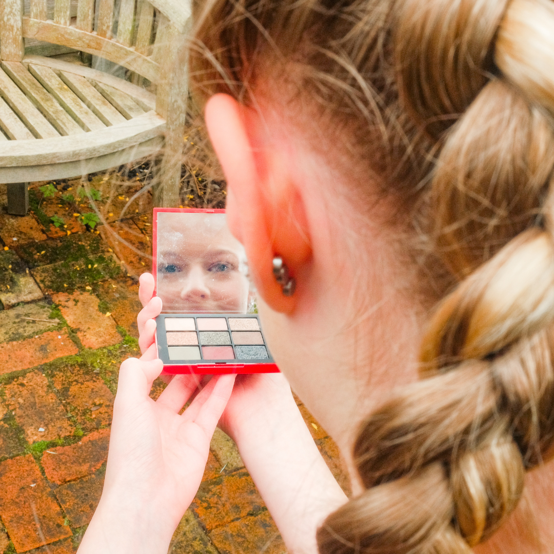 Teenage girl with braided hair holding an eyeshadow palette with a mirror, checking her reflection outdoors.