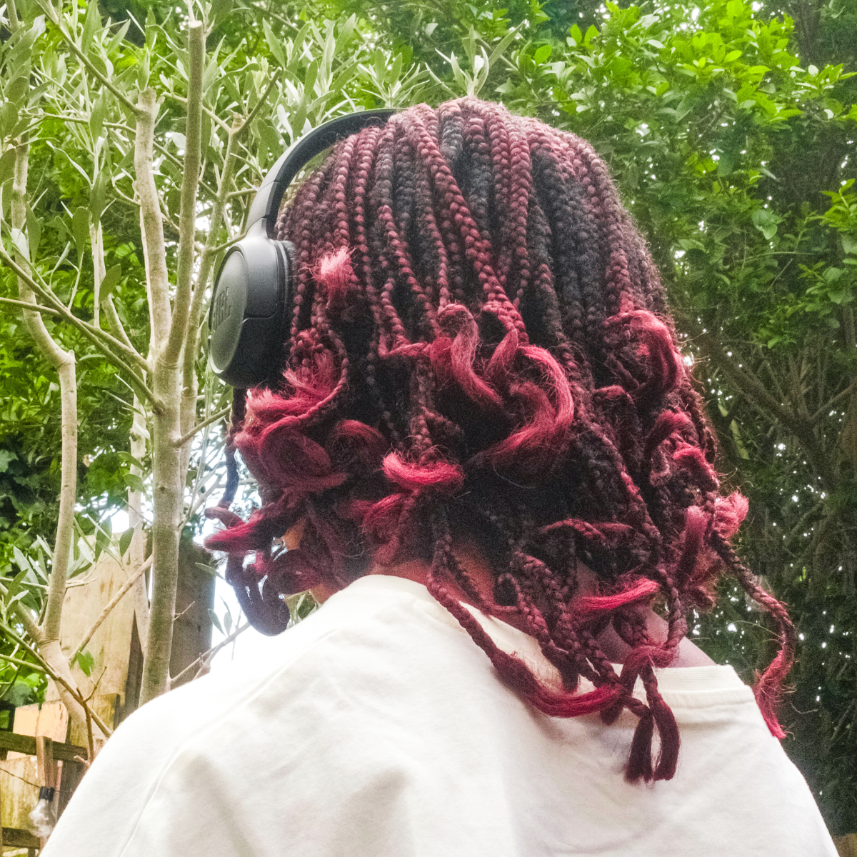 Back view of a teenager with braided red and dark hair wearing headphones outdoors among trees.