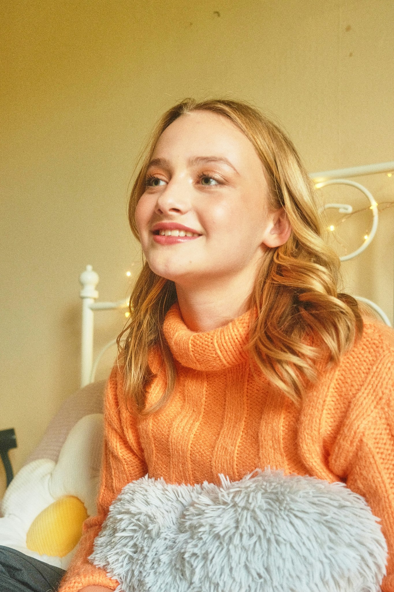 Teenage girl in an orange jumper smiling and looking to the side, with fairy lights in the background.