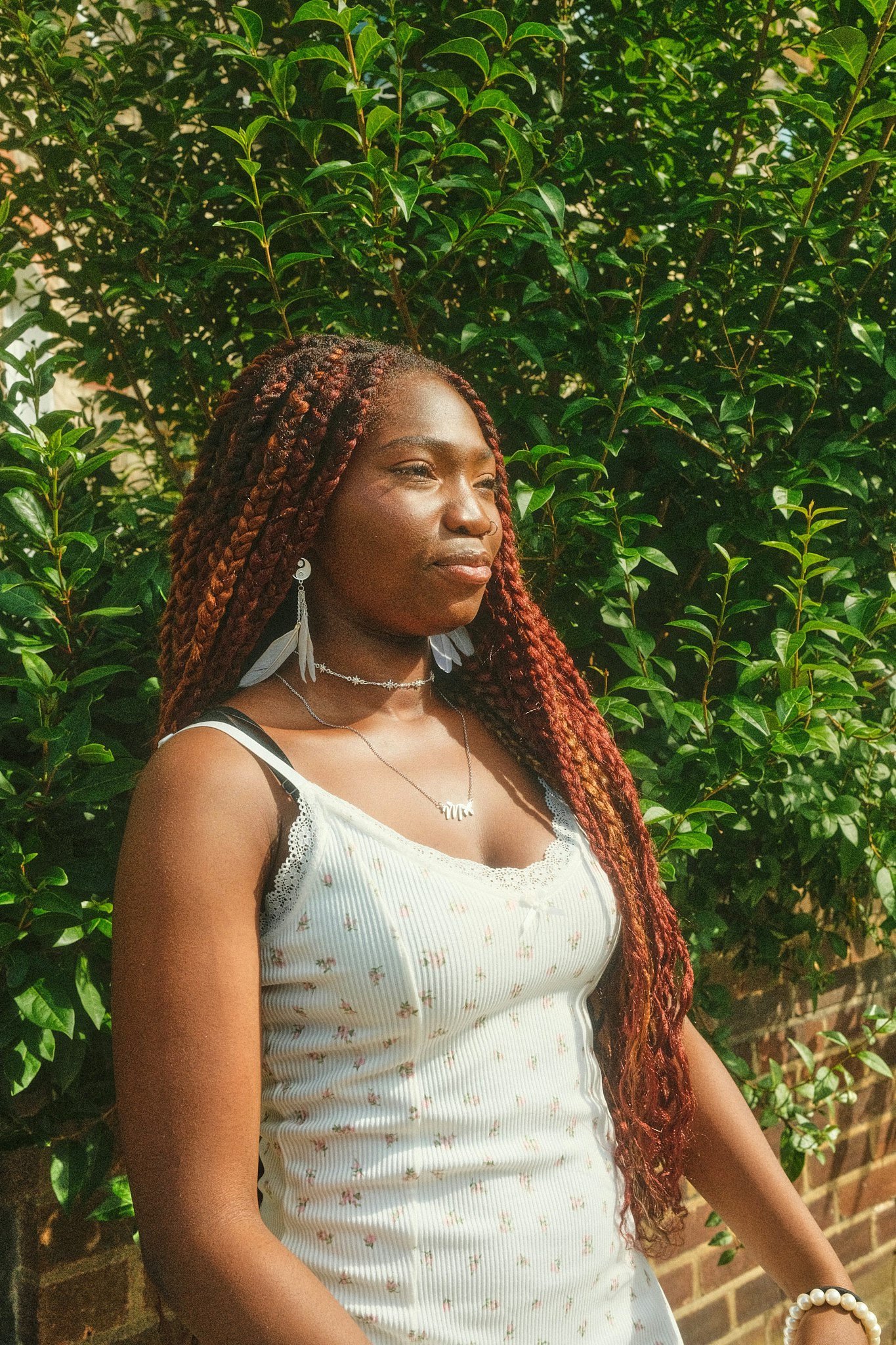 Teenage girl with long braided auburn hair looks off to the side outdoors, wearing a cream lace-trimmed top against a green leafy backdrop.