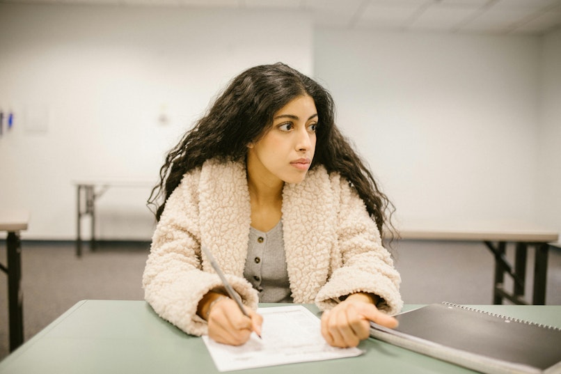 Teenage girl with curly hair wearing a cream jacket sits at a desk in a classroom, writing on paper with a pen while glancing to the side.