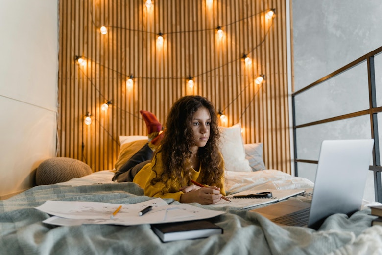 Teenage girl with curly hair in a yellow top lies on a bed doing homework with papers and a laptop, in front of a wall strung with warm fairy lights.
