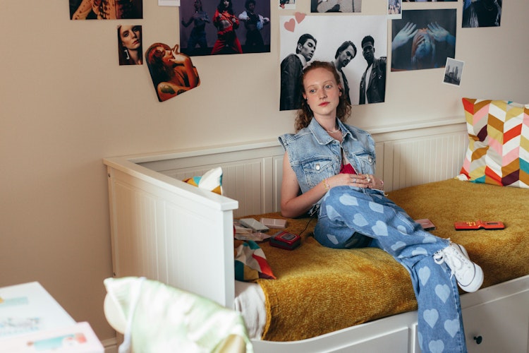 Teenage girl in a denim jacket and heart-print jeans sits on a bed in a messy, poster-covered bedroom with a colourful zigzag cushion.
