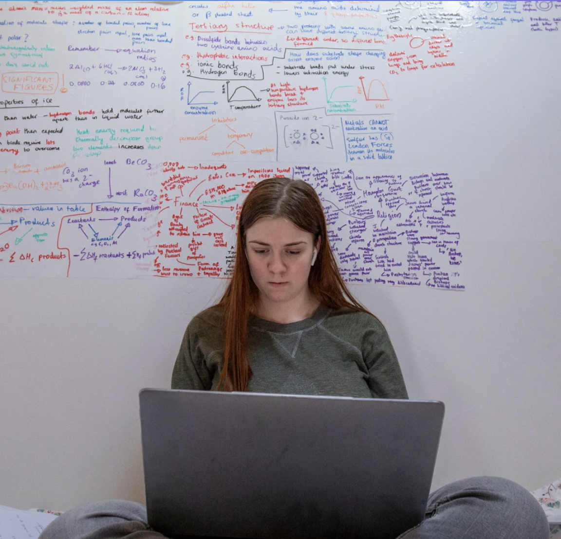Teenage girl with long red hair sits with a laptop in front of a whiteboard covered in colourful chemistry revision notes and diagrams.