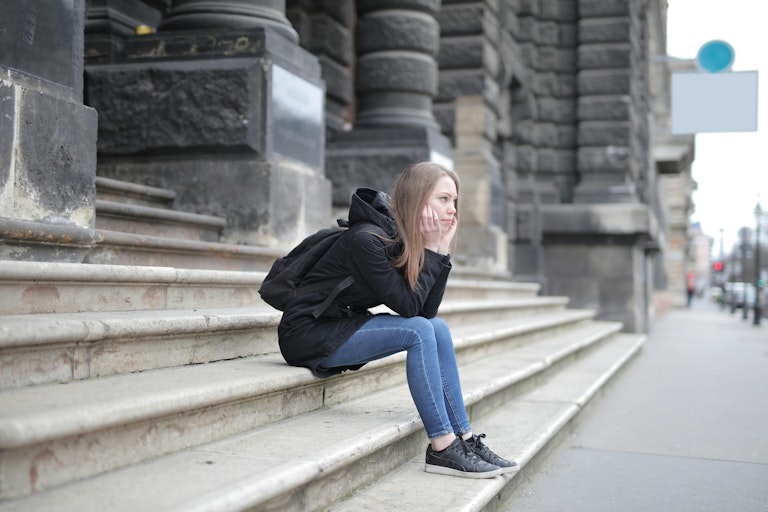 Teenage girl with a backpack sits alone on stone steps outside a large building, resting her chin on her hand and looking sideways with an unhappy expression.