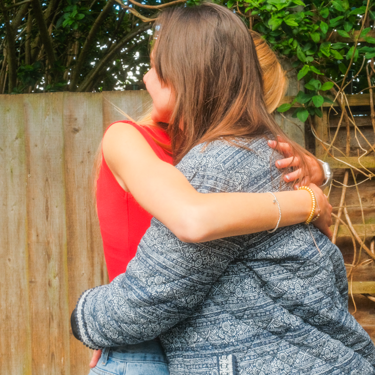 A mother and daughter hug each other tightly outdoors in front of a wooden fence and greenery, the daughter wearing a red top and the mother a patterned cardigan.