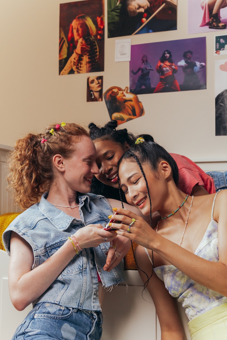 Three teenage girls with flower hair clips lean in close together laughing, in a bedroom with posters on the wall.