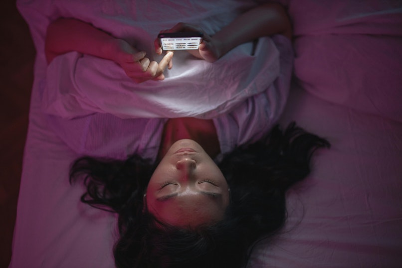 Teenage girl with dark hair lies in bed at night, eyes closed, holding a lit-up phone above her face in a dark, pink-lit room.