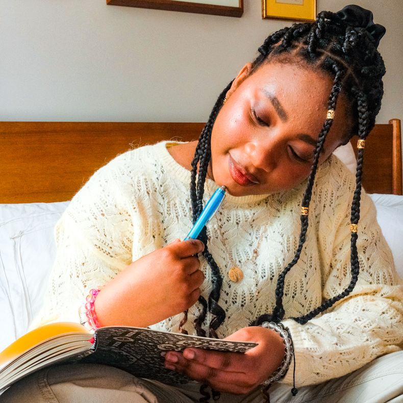 Teenage girl with braided hair and gold hair cuffs sits on a bed writing in a book with a blue pen, wearing a cream knit jumper.