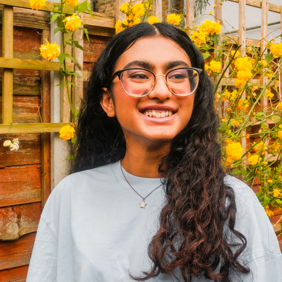 Teenage girl with glasses and curly hair smiles outdoors in front of yellow flowers and a wooden trellis.