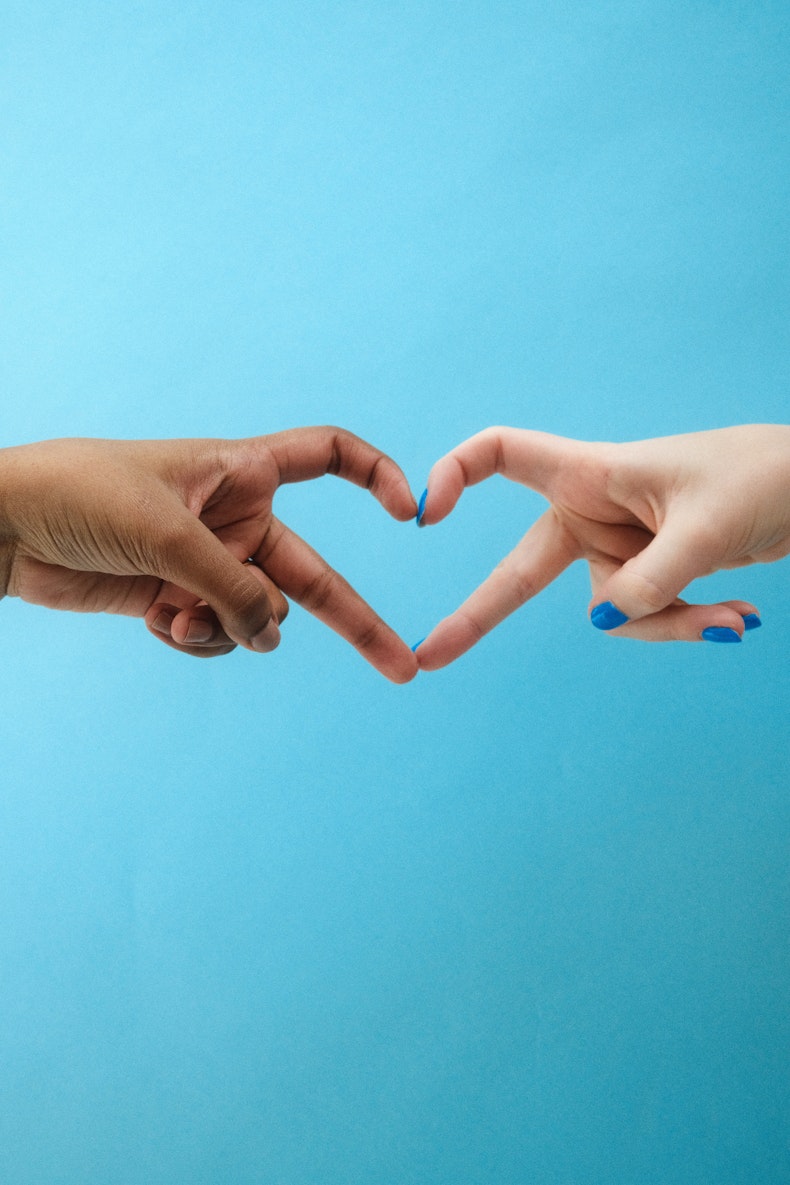 Two hands, one with darker skin and one with lighter skin and blue nail polish, form a heart shape together against a blue background.