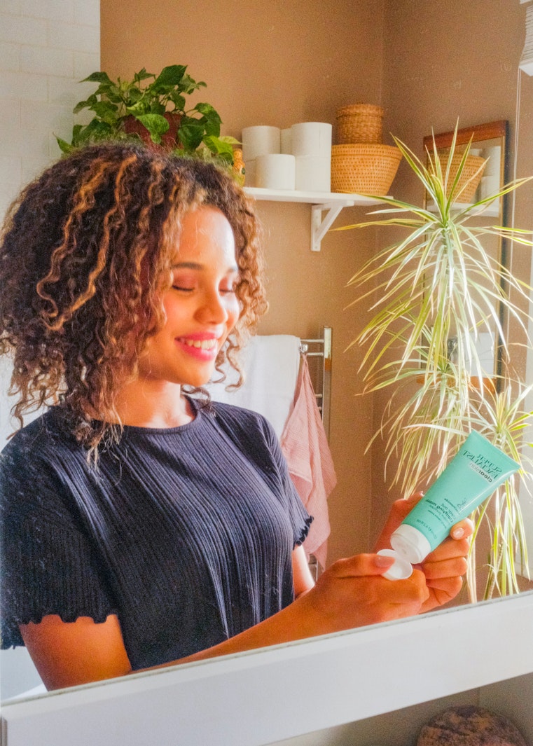 Teenage girl with curly hair smiles while holding a skincare tube in a bathroom with shelves and a plant.