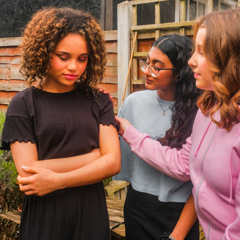 A teenage girl stands with her arms crossed looking downcast, while two other teenage girls stand close together behind her, one resting a hand on her shoulder, outdoors in a garden.