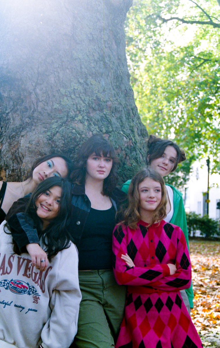 A group of five teenage girls in autumn outfits stand and lean against a large tree trunk in a park with fallen leaves.
