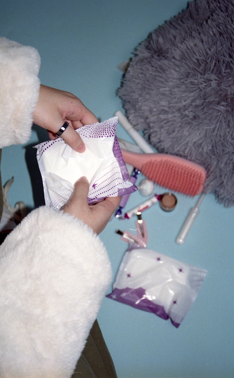 Hands in a fluffy white coat hold open a packet of period pads, with a hairbrush and a bag of more pads on a bed in the background.