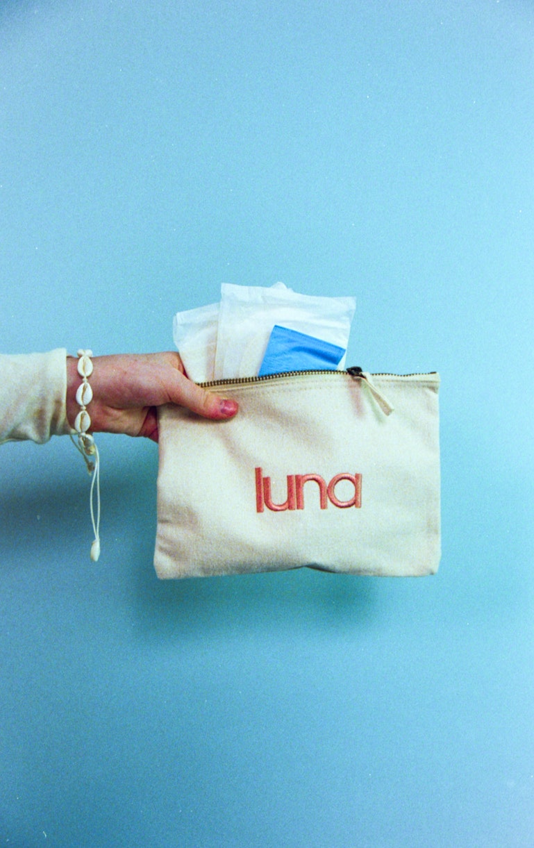 A hand holds up a cream luna pouch containing period products against a blue background.
