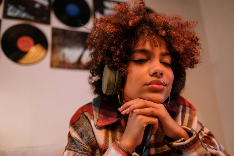 Teenage girl with curly auburn hair wears headphones and rests with her eyes closed and chin on her hands, in front of a wall decorated with vinyl records.