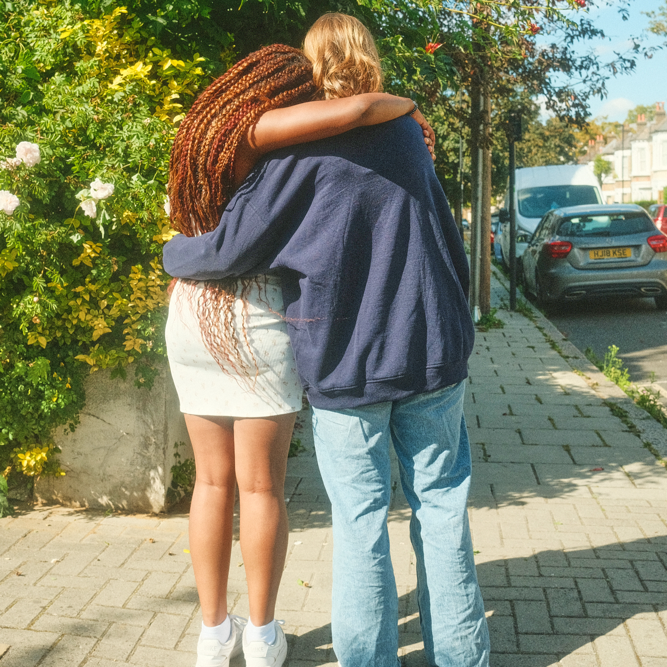 Two teenage girls, one with braided hair and one with blonde hair, hug each other with their arms around each other's shoulders, walking down a residential street.