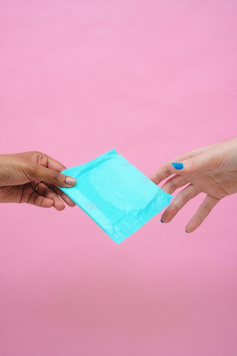 One hand passes a wrapped period pad to another outstretched hand against a pink background.