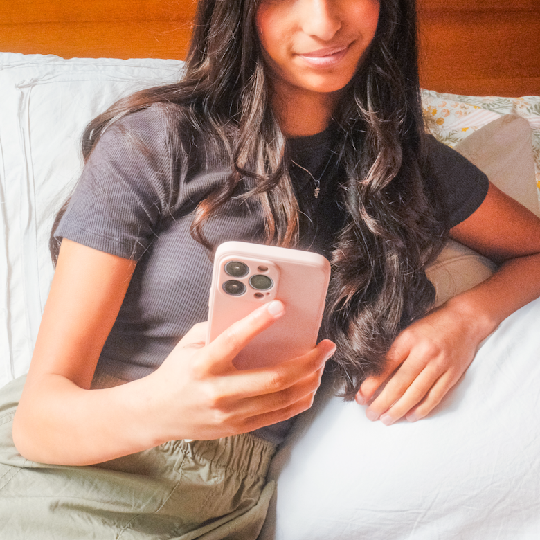 Teenage girl with long curly hair lies on a bed holding up a phone, smiling slightly.