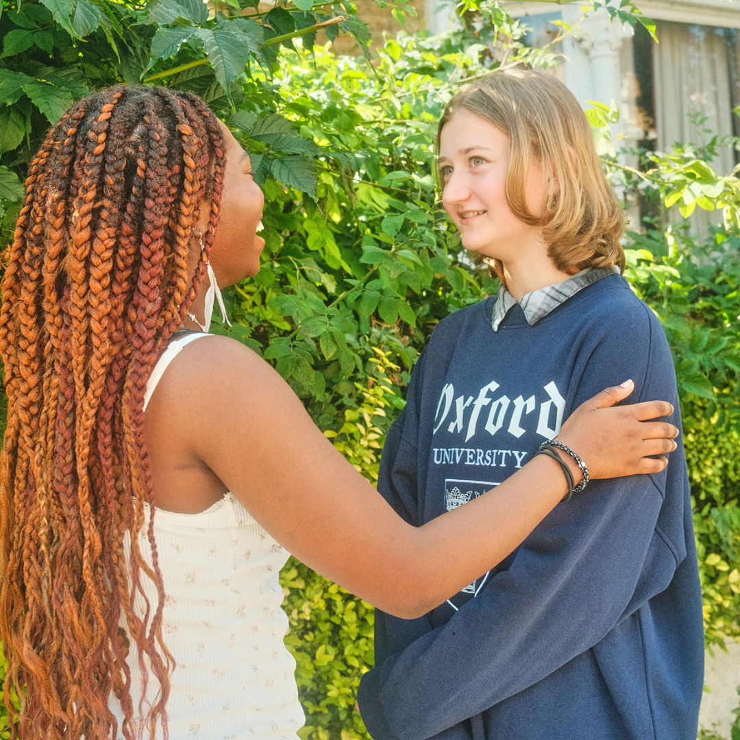 Two teenage girls smiling at each other warmly outdoors, one with long braided hair, the other wearing a navy Oxford University sweatshirt.