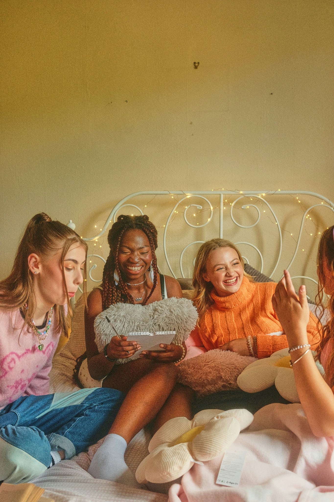 Four teenage girls sitting together on a bed laughing in a cosy bedroom with fairy lights.