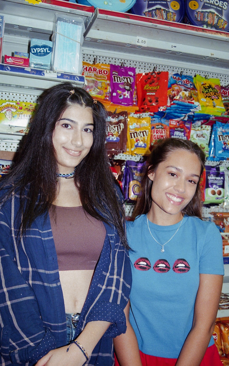 Two teenage girls smiling together in a sweet shop with brightly packaged sweets on the shelves behind them.
