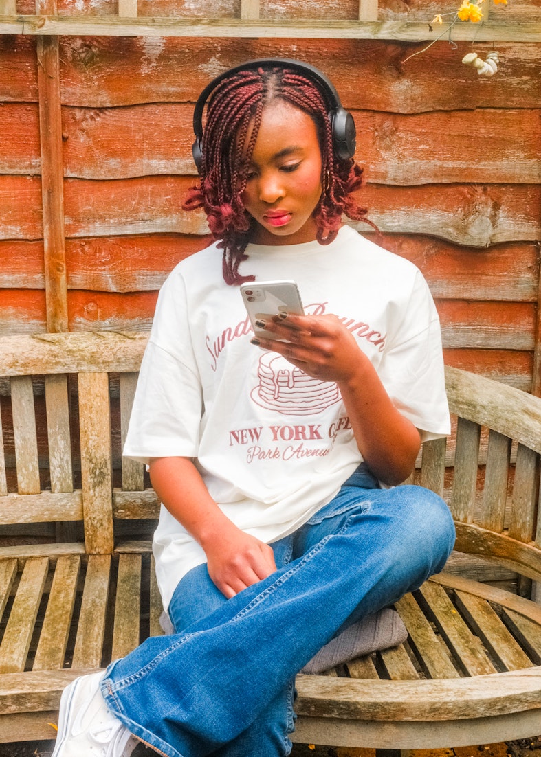 Teenage girl with red braided hair wearing headphones sits on a wooden bench looking at her phone.