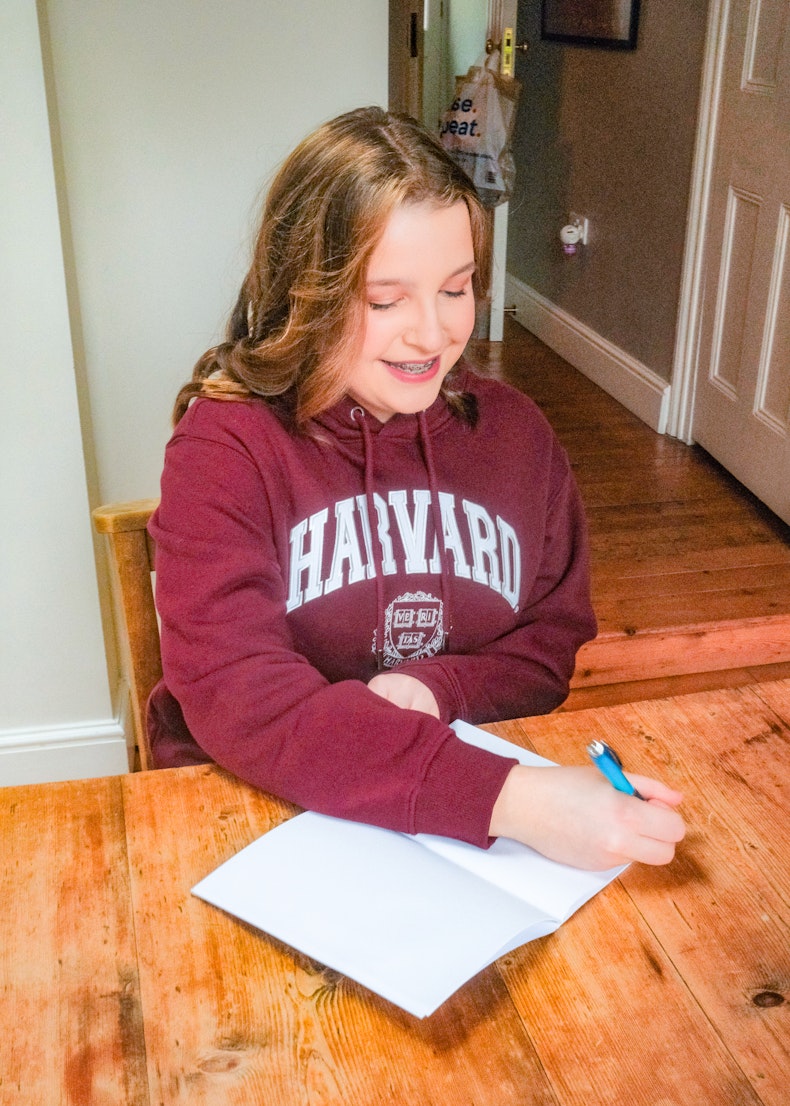 Teenage girl in a Harvard hoodie smiles while writing in a notebook at a wooden table.