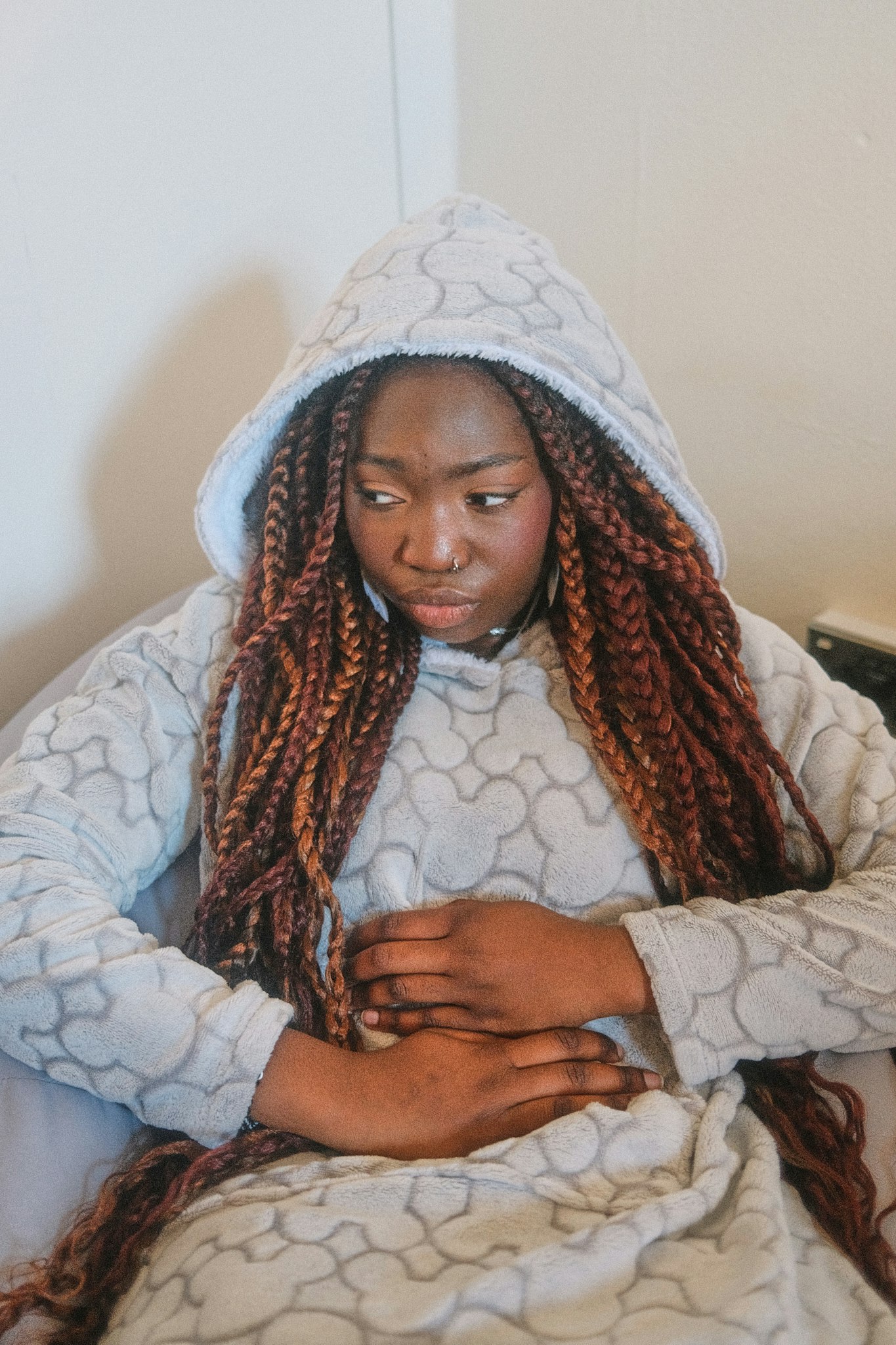 Teenage girl with braided hair sits hunched forward holding her stomach, wearing a hooded fleece.