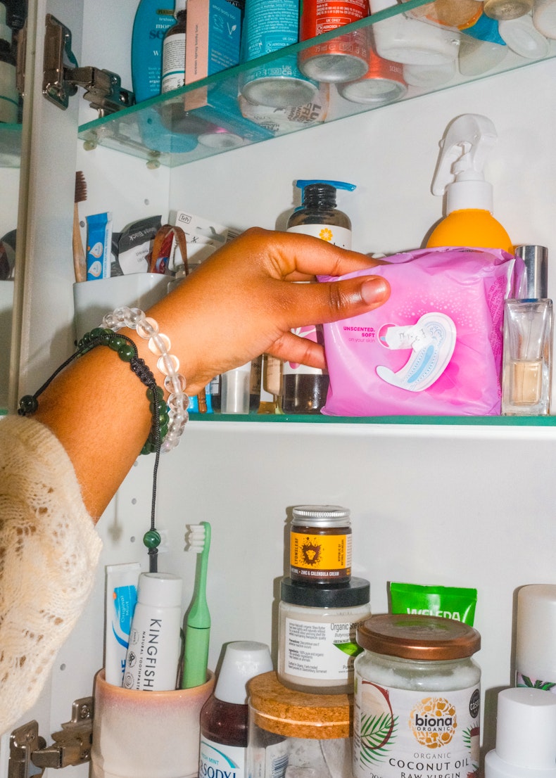 Hand reaching into a bathroom cabinet to take out a pink packet of period pads, surrounded by toiletries.