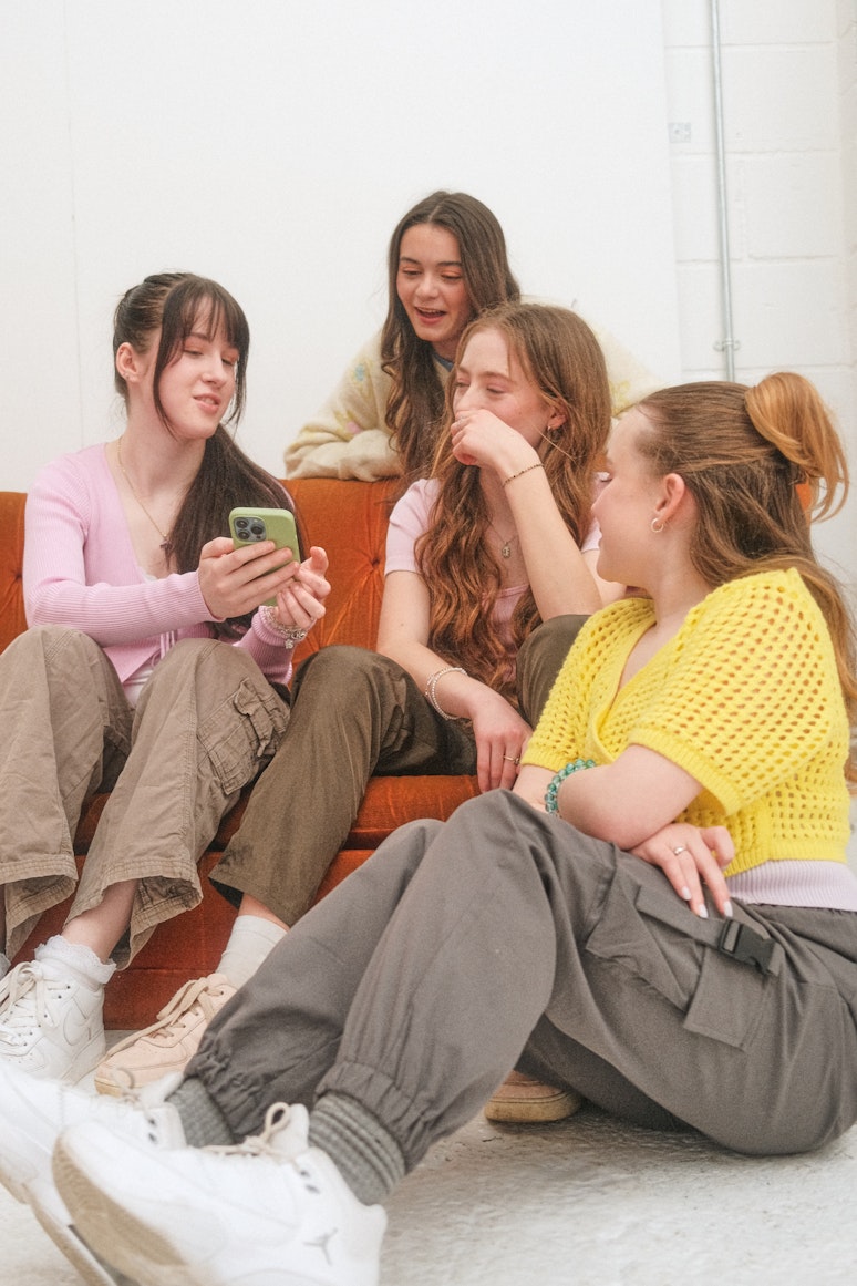 Four teenage girls sitting together on a sofa, laughing and looking at a phone screen.