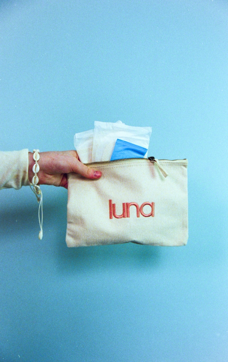 Hand holding a cream luna pouch with period pads tucked inside, against a blue background.