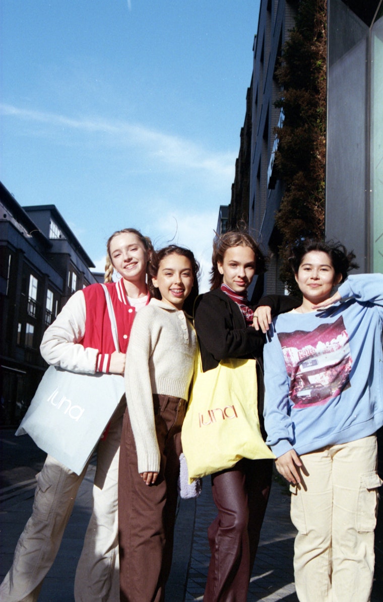 Four teenage girls smiling together on a city street, two holding luna tote bags.