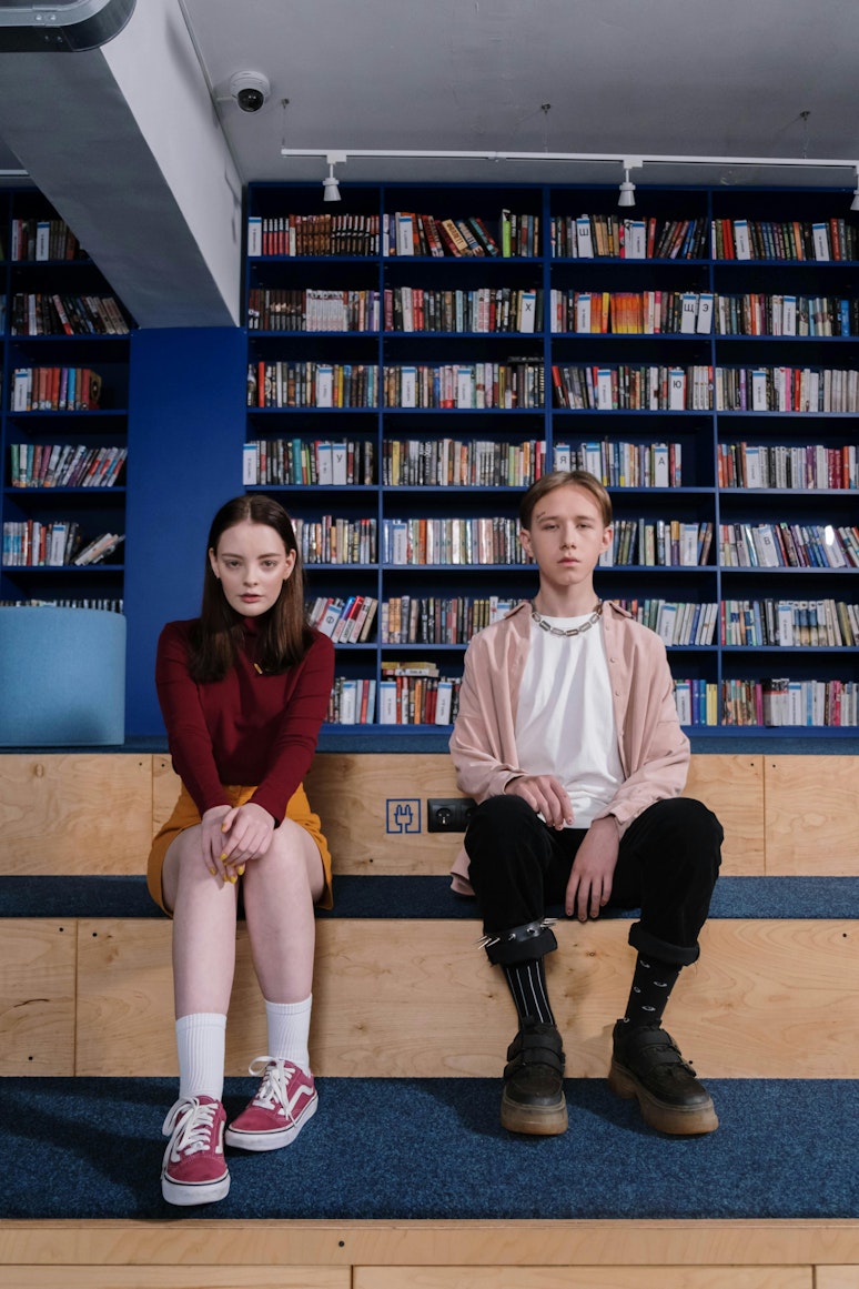 A teenage girl and boy sit apart on a bench in a library, both with serious expressions.