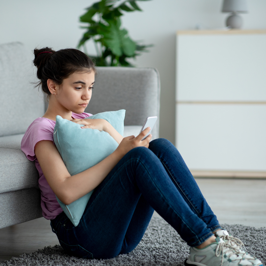 Teenage girl sitting on the floor hugging a cushion while looking at her phone with a concerned expression.