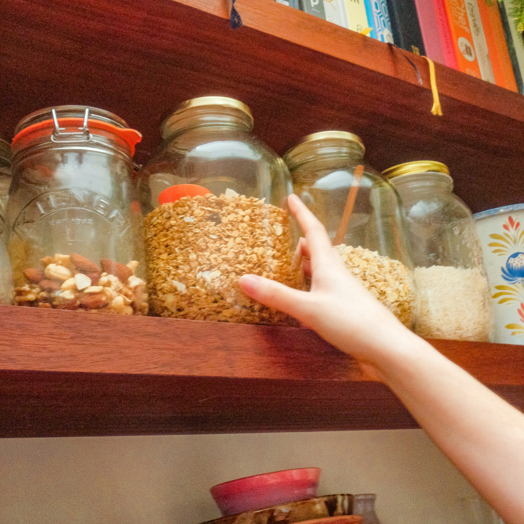 A teen's hand reaches up to a jar of granola on a shelf of glass jars filled with nuts, oats and rice.