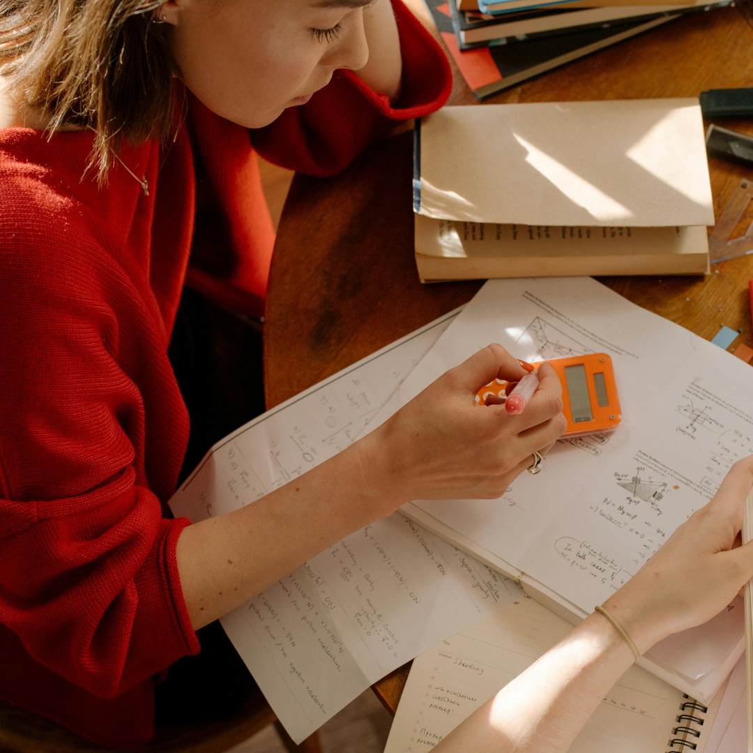 Two teenage girls studying together at a table covered in notes and textbooks, one using a calculator.